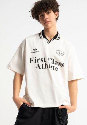 Młoda osoba z kręconymi włosami, ubrana w białą, oversize'ową koszulkę polo z napisem "First Class Athlete" oraz czarne szorty z łańcuchami.
