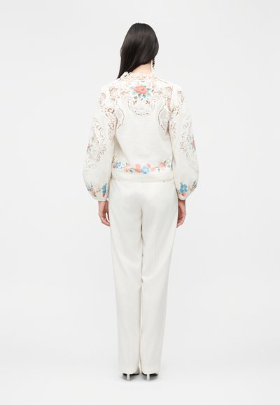Γυναίκα με μακριά σκούρα μαλλιά στέκεται με την πλάτη, φοράει λευκό παντελόνι και λευκό πουκάμισο με λουλουδάτη κεντητή διακόσμηση και δαντελένιες λεπτομέρειες στην πλάτη και στα μανίκια.
