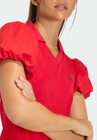 Červené tričko s nafouklými rukávy, výstřihem do V a detaily s logem. Hladká textura a lesklý povrch látky.