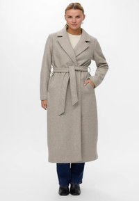 Sieviete, tērpusies garā beidžā jostā ietīktā vilnas mētelī, virs krēmkrāsas džempera, zilajiem džinsiem un melnajām potītēm, stāv ar vienu roku kabatā.