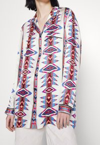 Chemise en soie avec une base blanche ornée de motifs géométriques audacieux en rouge, bleu et noir. Comprend une poche avant et un design à boutonnière.