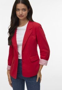 Blazer rouge avec revers crantés, deux poches frontales et détails rayés sur les poignets. Associé à une chemise blanche et un jean en denim.