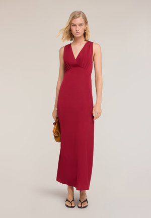 Junge Frau trägt ärmelloses, tiefrotes Maxikleid mit V-Ausschnitt, hält eine braune Clutch und steht vor schlichtem hellem Hintergrund.