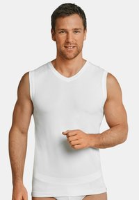 Débardeur sans manche blanc en V, fabriqué en tissu lisse, au design ajusté, mettant en valeur une silhouette épurée. Le mannequin pose avec une expression neutre.
