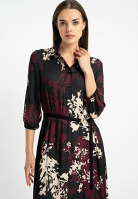Kobieta w czarnej sukience w burgundowe i beżowe kwiaty, z rękawami do trzech ćwiartek, z guziczkowym kołnierzykiem i cienkim czarnym paskiem w talii.