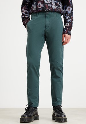 Mężczyzna w turkusowych obcisłych spodniach, kwiecistej koszuli z długim rękawem i czarnych butach na sznurowadła z grubą podeszwą stoi na jasnej podłodze.