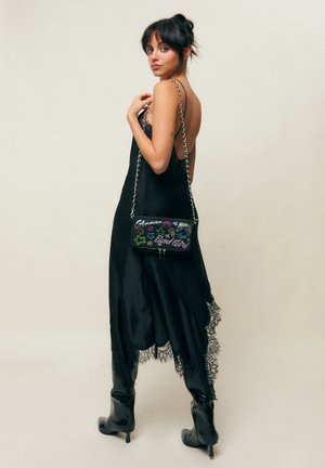 Frau trägt ein schwarzes Negligé-Kleid und Stiefel mit Absatz, hält eine kleine schwarze Umhängetasche mit bunten Schriftzügen und Sternmotiven.