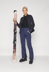 Skidutstyr består av en svart jakke med blå detaljer, høytrukne marineblå bukser, svarte skistøvler og en brun ski med bindinger.