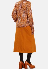 Blusa a maniche lunghe con motivo floreale arancione, con polsini arricciati e pieghe sul retro; abbinata a una gonna di jeans color ruggine fino al ginocchio e scarpe con zeppa.