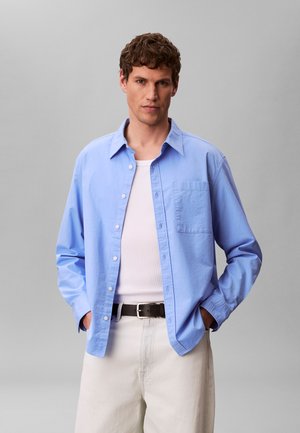 Mann trägt hellblaues Hemd mit Knöpfen über weißem Tanktop, beige Hose und schwarzen Gürtel, steht mit Händen in den Taschen vor grauem Hintergrund.