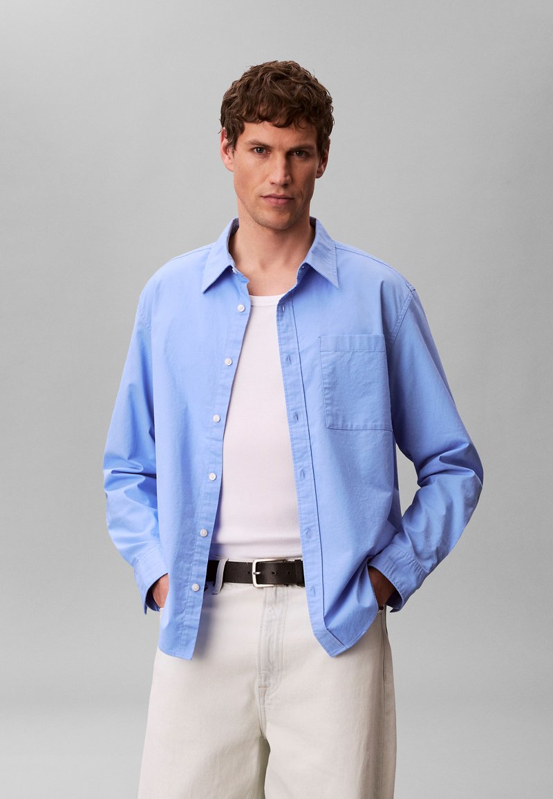 Homme portant une chemise bleu clair boutonnée sur un débardeur blanc avec un pantalon beige et une ceinture noire, debout les mains dans les poches sur fond gris.