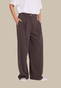 Vidde bukser i mørkebrunt stof med sidelæg, med knaplukning foran og dype lommer. Parret med hvite joggesko.