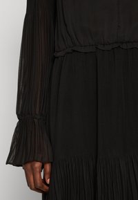 Svart klänning med veckade, genomskinliga ärmar och rynkad midja. Texturerat tyg med volangkant och detaljerade ärmslut.