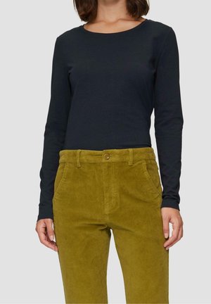 Kobieta ubrana w prostą czarną bluzkę z długim rękawem, wpuszczoną do wysokich sztruksowych spodni w kolorze musztardowym z kieszeniami i zapięciem na guziki.