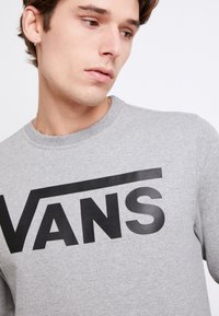 Sudadera gris con un destacado logo negro de "VANS". Presenta un cuello redondo y una tela suave y texturizada. Diseño sencillo, estilo casual.