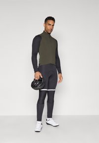 Fietsoutfit met een groene ritsvest en zwarte lange tights met witte strepen. Het model houdt een zwarte helm vast en draagt witte fietsschoenen.