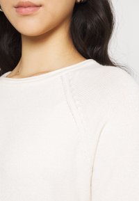 Pull crème montré en gros plan, avec un col rond, une texture tricot côtelée et des coutures d'épaule subtiles. Matériau doux et léger.