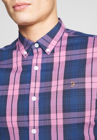 Skjorta med knappar framtill som har ett blått och rosa rutmönster. Tillverkad av tyg med en slät yta, inkluderar en liten broderad logga på bröstet.