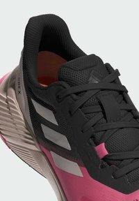 Zapatilla deportiva negra con parte superior de malla, acentos rosas y rayas grises. Presenta cordones texturizados y una suela acolchada para mayor soporte.