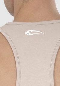 Licht beige tanktop met een ronde hals, gemaakt van een gladde stof. Voorzien van een wit logo op de bovenrug.