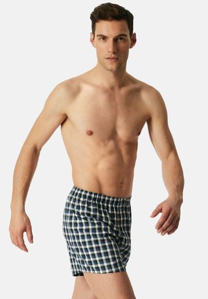 Blaue und weiße karierte Baumwollshorts mit elastischem Bund, mit lockerer Passform und einem Saum, der oberhalb des Knies endet, getragen von einem Modell.