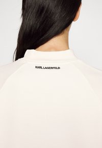 Dlouhé černé vlasy přes záda osoby, která má na sobě bílé tričko se vyšívaným logem "KARL LAGERFELD" na šíji.