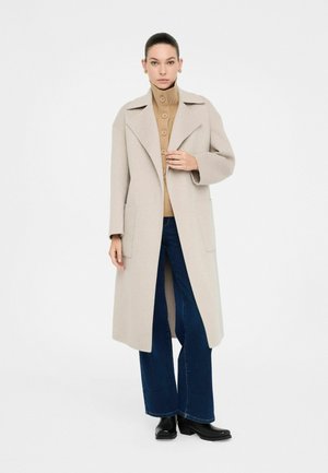 Cappotto in misto lana beige con colletto a scialle, tasche frontali e silhouette dritta, abbinato a un pullover button-up color sabbia e jeans scuri.