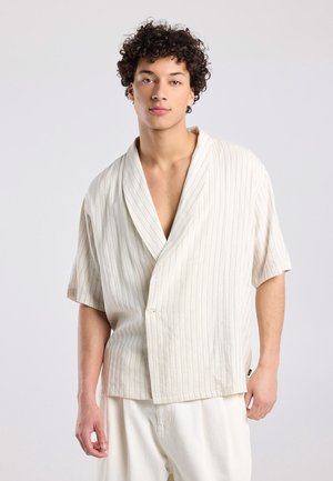 Jonge man met krullend haar, gekleed in een losse, kortärmelige, lichtbeige gestreepte blouse en lichtgekleurde broek, tegen een effen achtergrond.