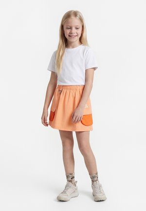 Jong meisje met lang blond haar draagt een wit T-shirt, perzikkleurige rok met oranje zakken, sokken met patroon en beige sneakers, glimlachend.