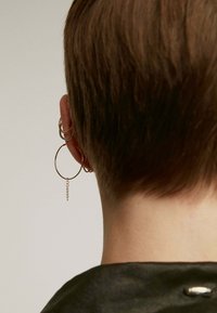 Pendientes de aro en tono dorado con un acento de cadena, exhibidos contra un cabello corto castaño, mostrando un cuello angular y ropa oscura.