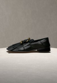 Μαύρο δερμάτινο loafer με χρυσές μεταλλικές λεπτομέρειες και φούντες, εκτεθειμένο σε ανοιχτή επιφάνεια σε γκρι φόντο.