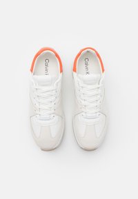 Baskets blanches Calvin Klein avec accents orange au talon, design à lacets et semelles intérieures marquées, vues de dessus sur un fond uni.
