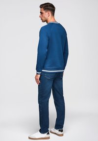 Mladý muž otočený vlevo, oblečený v modré mikině s bílým lemováním, tmavomodrých džínách a bílých teniskách s hnědými podrážkami.