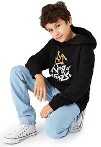 Svart hoodie med gyllene krona och vit text "King of Fools" på framsidan, kombinerad med ljusblå jeans och vita sneakers med svarta accenter.