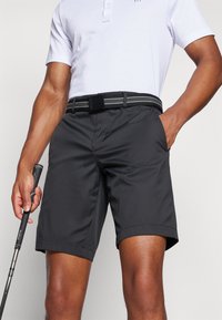 Calções de golfe pretos com frente lisa, bolsos laterais e passantes para cinto. Feitos de um material leve e texturizado. Usados com uma camisola polo branca.