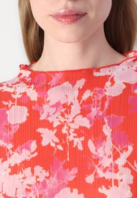 Blusa floreale rossa con motivi rosa e bianchi, texture plissettata e scollo rotondo con dettaglio a fiocco sul retro.