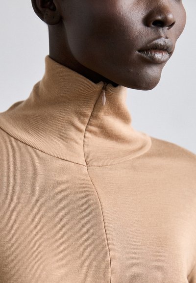 Tan ribbed turtleneck sweater with a half-zip closure. Features a smooth texture and fitted shape with subtle seam detailing.