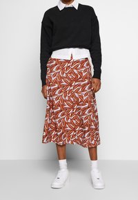 Felpa nera cropped indossata sopra una camicia bianca con colletto, abbinata a una gonna di lunghezza media con un motivo di foglie marroni e bianche, indossata con sneaker bianche.