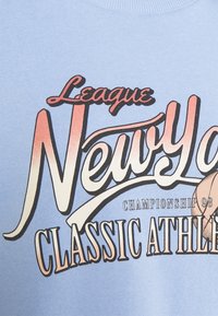 Ljusblå sweatshirt med fet grafisk text "League New York Classic Athletic Championship 98" och en basketillustration.