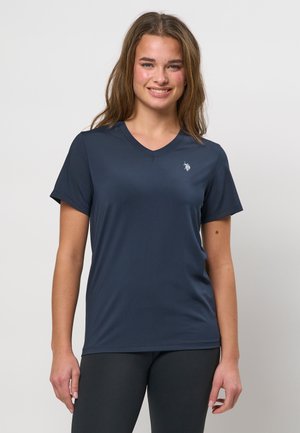 Noor naine, kellel on pikad juuksed, seljas tumesinine V-kaelusega lühikeste varrukatega särk ja mustad retuusid, naeratab ja seisab ühevärvilise taustaga.