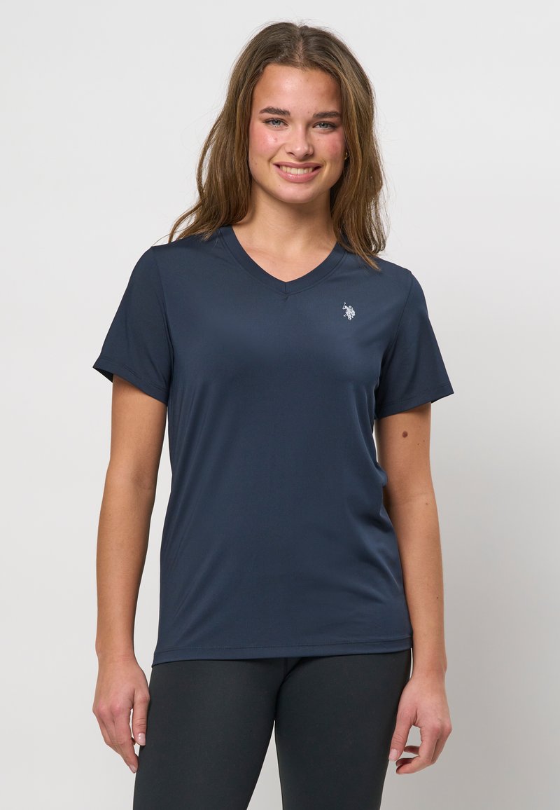 Młoda kobieta o długich włosach, ubrana w granatową koszulkę z dekoltem w serek i krótkim rękawem oraz czarne legginsy, uśmiechająca się i stojąca na jednolitym tle.