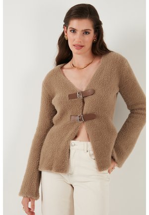 Tan fuzzy cardigan with a V-neck, featuring two brown strap buckles for closure. Long sleeves and a relaxed fit.