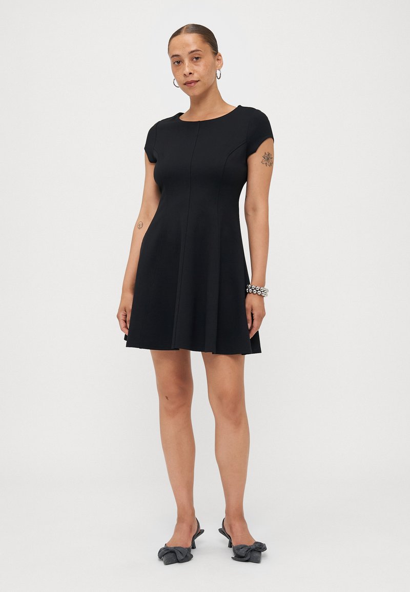 Robe noire à manches courtes avec un corsage ajusté et une jupe évasée, fabriquée en tissu lisse ; accompagnée de chaussures noires ornées d'un nœud.