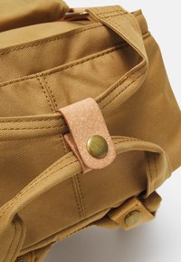 Beige stoffryggsekk hjørne med synlige sømmer, en lærrem-lukking og messingknapper for sikker festing.