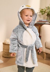 Peuter in een grijs-witte pluche pinguïn ochtendjas met capuchon binnen, staand naast een rotan mand en een fauteuil in een woonkamer.