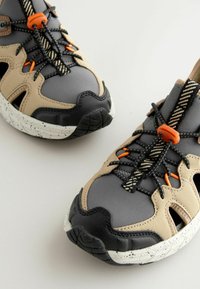 Baskets de sport avec une tige en synthétique gris, beige et noir, des panneaux en maille ouverte, des accents orange et une semelle en caoutchouc texturé avec des éclats.