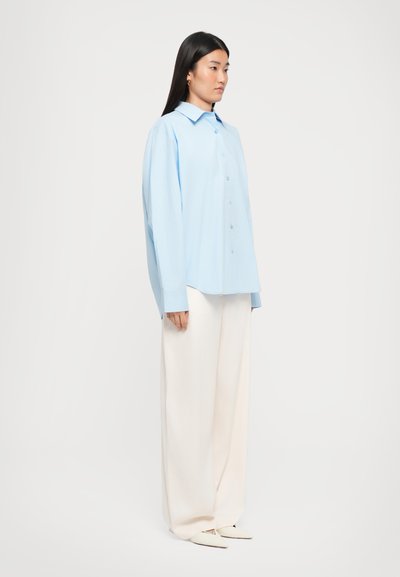 Hellblaues Button-Up-Hemd mit spitzem Kragen, lässiger Passform und langen Ärmeln, kombiniert mit cremefarbenen, weit geschnittenen Hosen und spitzen weißen Schuhen.