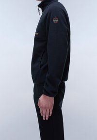 Czarna polarowa kurtka z wysokim kołnierzem, zamykaną na zamek kieszenią na froncie i naszywką z logo na górnej części rękawa. Nosi się z czarnymi spodniami; gładka faktura.