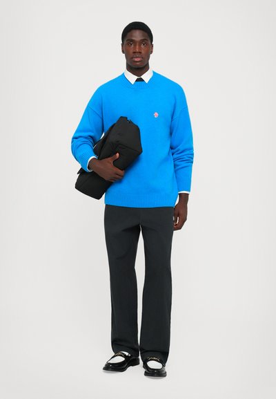 Blauer Strickpullover, schwarze Hose und schwarze Schuhe mit weißen Akzenten. Hält eine schwarze Tasche, mit einem unter dem Pullover sichtbaren weißen Kragen.