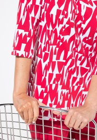Rood en witte top met een patroon en korte mouwen, met abstracte vormen. Handen die een handvat van een winkelwagentje vasthouden.